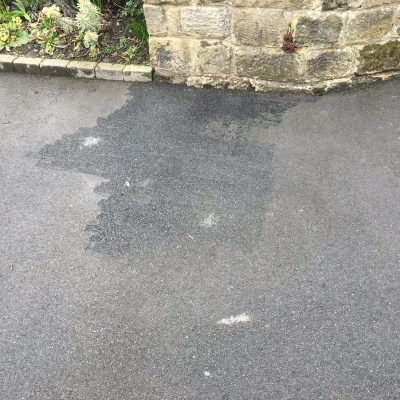 An initial cleaning test patch on a weathered tarmac driveway beside a stone garden wall.