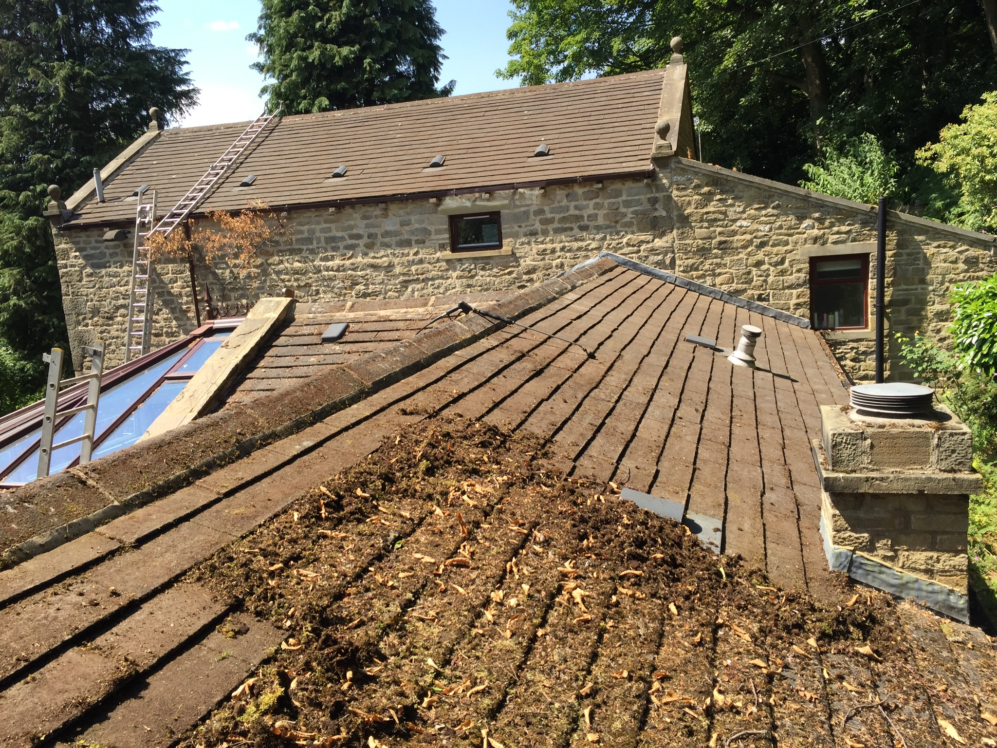 Loose moss and organic debris gathered on a large farm house concrete-tiled roof during manual removal, with cleared roof sections visible behind.