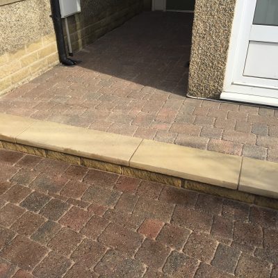 Freshly sealed block paving around a front entrance, showing a richer red-and-charcoal finish beside the low stone step.
