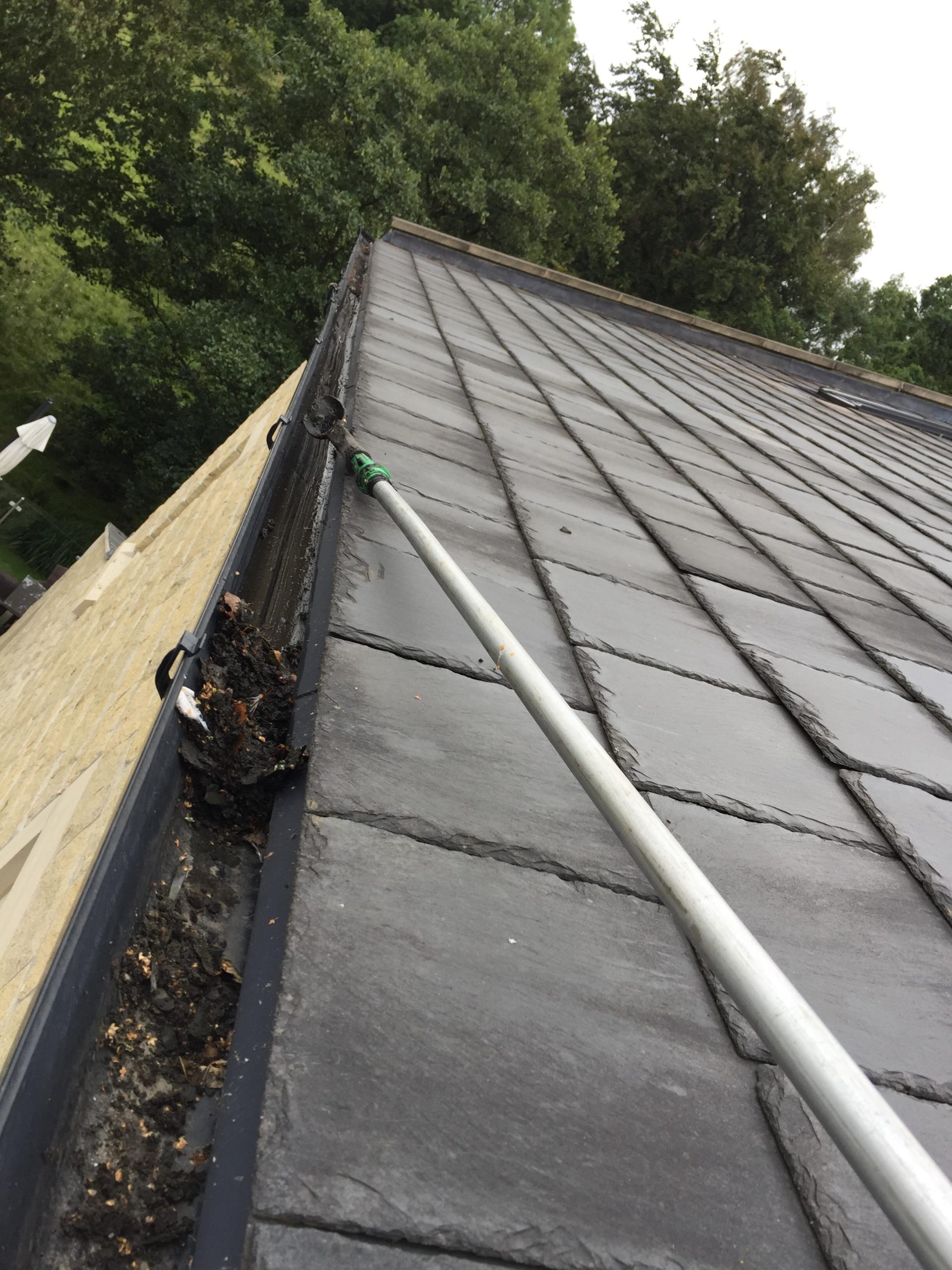 long-handled gutter scoop removing compacted leaves and mud from the gutter beside a natural slate roof.
