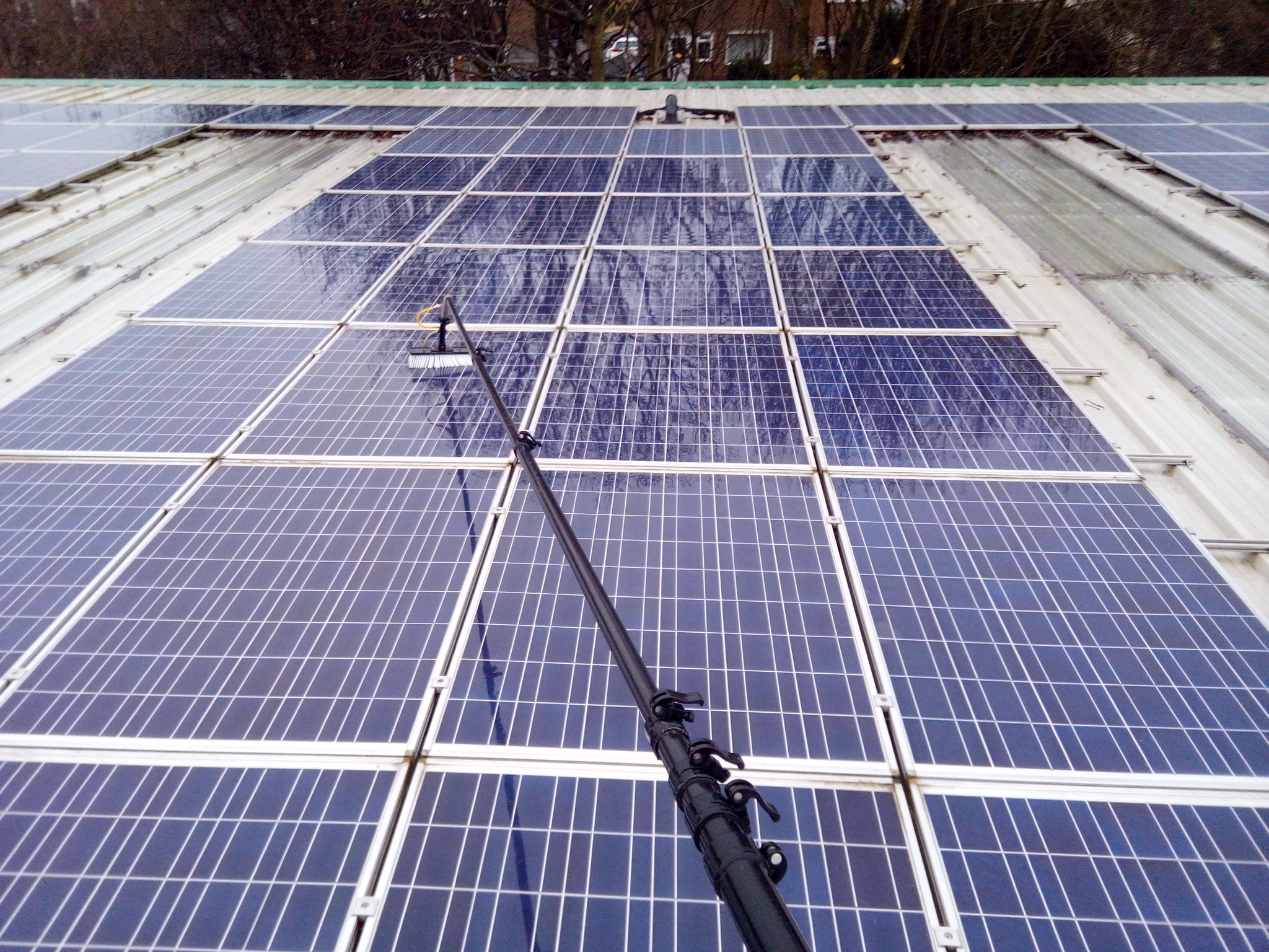 A water-fed pole brush cleaning a large solar panel array mounted on a profiled metal roof.