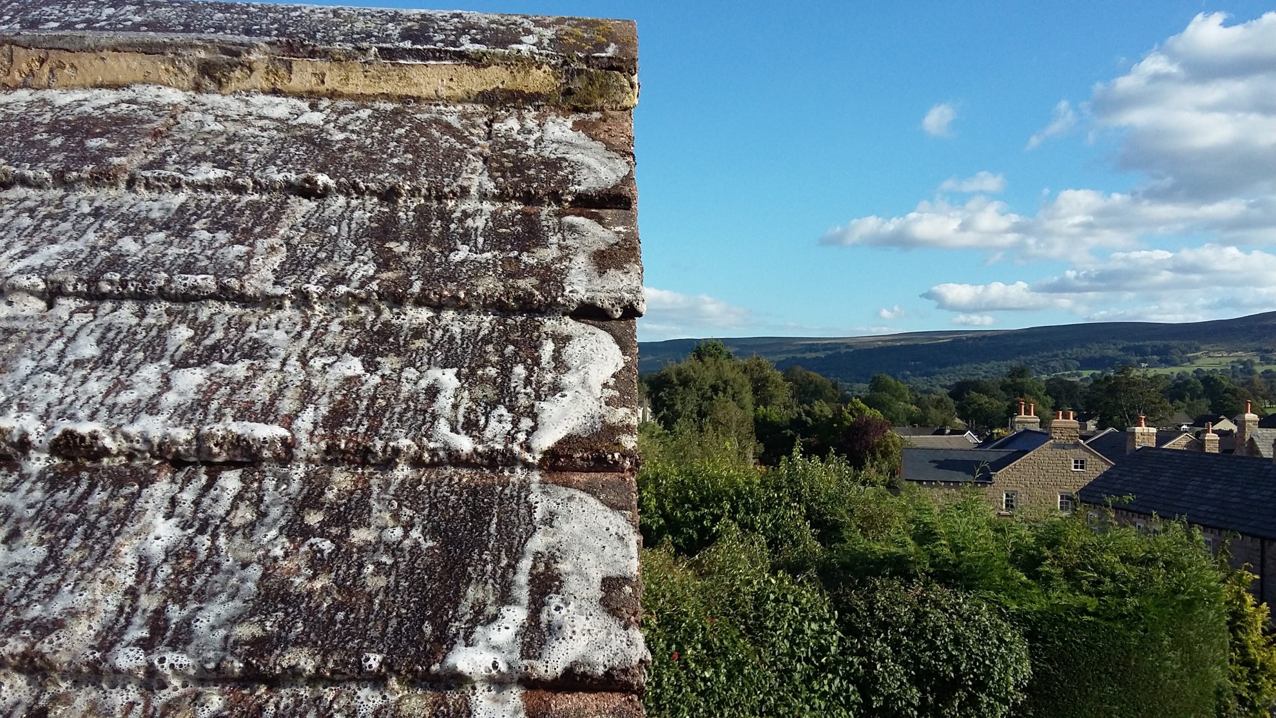Foaming roof-cleaning treatment covering dark flat-profile tiles, with Otley’s stone houses and surrounding hills visible beyond.