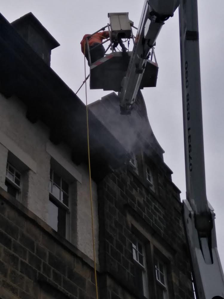 An operative cleaning gutters at roof level from a cherry picker beside a multi-storey stone building.