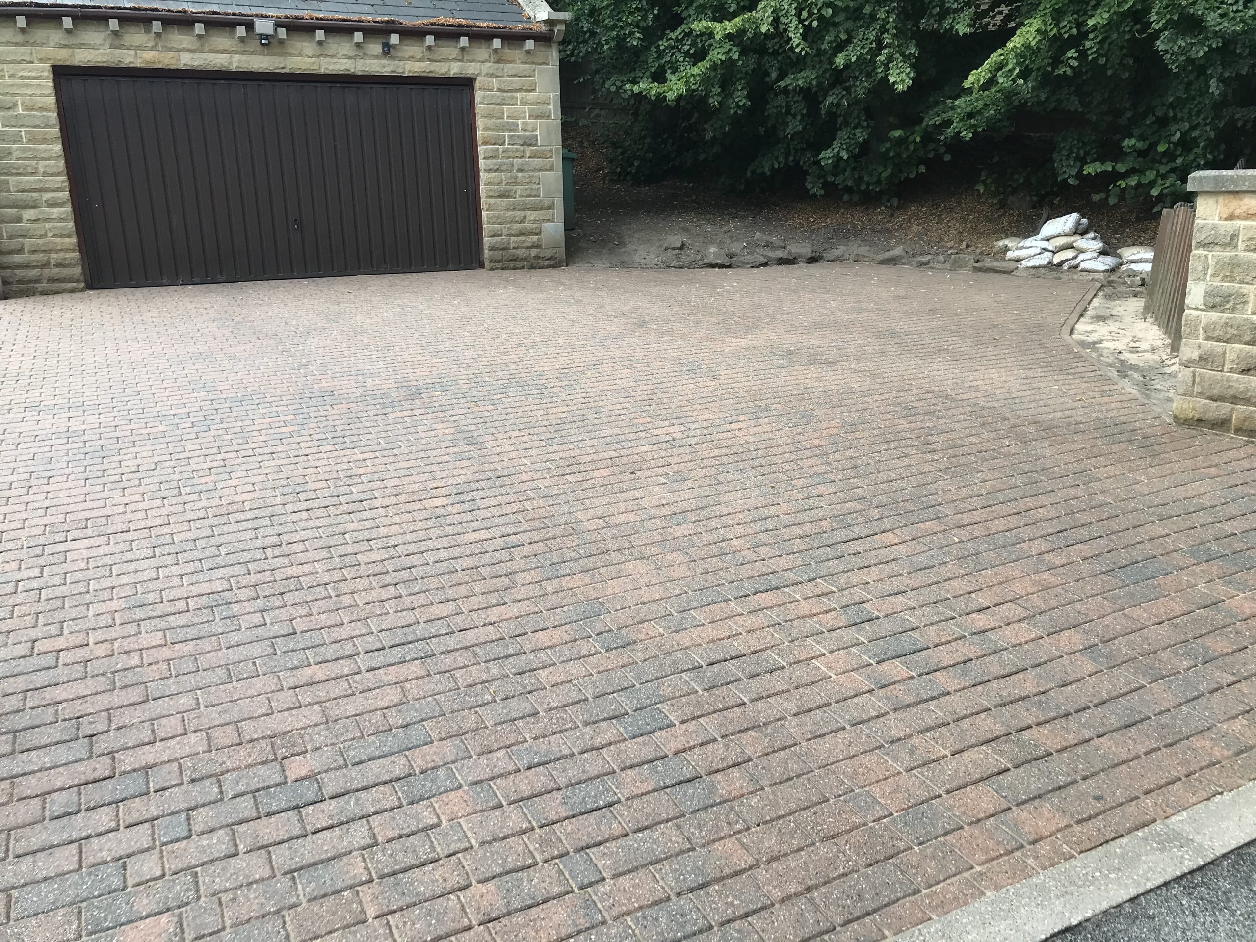 A wide, clean residential driveway with red-brown and charcoal concrete blocks laid in a herringbone pattern outside a stone-built garage.