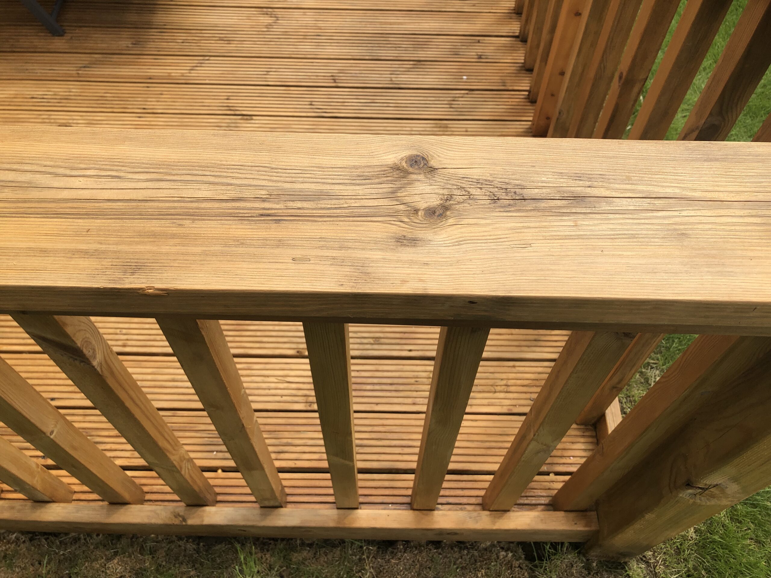 Close-up of a stained timber deck balustrade above grooved boards, showing a warm golden-brown finish with the grain and knots still visible.