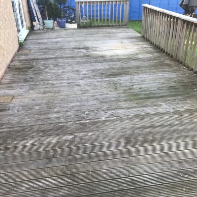 Large grooved timber deck beside a brick house, greyed by weathering and marked with green algae before cleaning and staining.